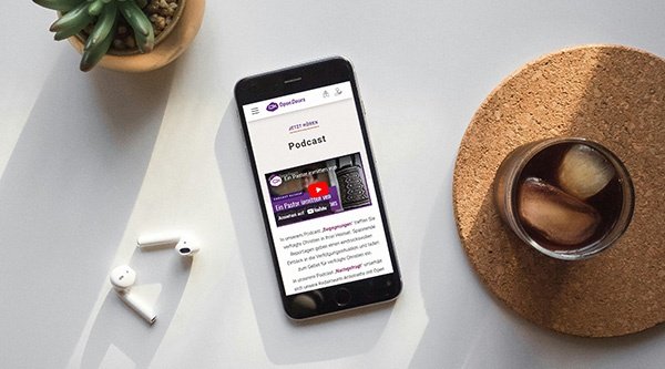 Ein Smarthphone liegt zwischen einer Tasse Tee und einer Sukkulente auf einem Tisch. Daneben liegen Bluetooth-Kopfhörer.
