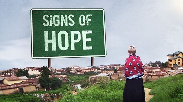 Eine Frau steht auf einem Feldweg vor einem nigerianischen Dorf. Im Hintergrund wurde ein großes Schild in das Bild eingefügt. Auf diesem steht in Capital Lettern der Schriftzug SIGNS OF HOPE.
