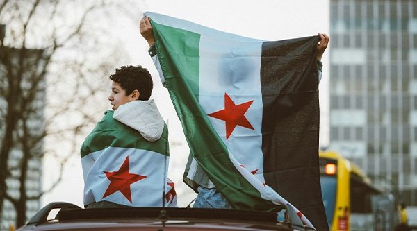 Stadtbild in Syrien. Zwei Personen sitzen eingehüllt in den Revolutionsflaggen Syriens auf einer Mauer.