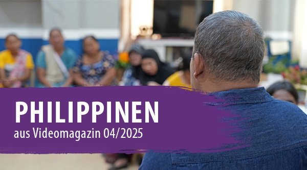 Hinterkopf eines Mannes mit blauem Shirt Im Hintergrund sitzen Menschen im Halbkreis die nur unscharf zu sehen sind