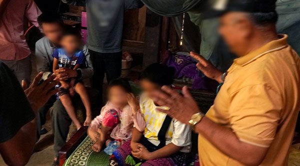 Kinder und Erwachsenen sitzen und ein Mann mit gelben Shirt hat seine Hände im Gebet ausgestreckt über die Kinder