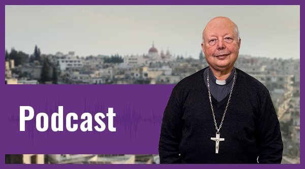 Ein Pastor in einem schwarzen Gewand und einer goldenen Kette mit Kreuz steht in Syrien vor einer Stadt.