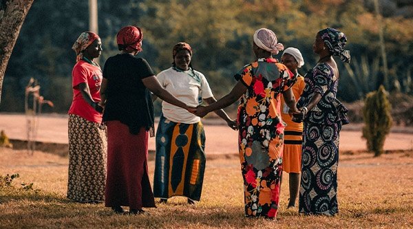 Eine Gruppe afrikanischer Frauen steht auf einer Wiese im Kreis und hält sich an den Händen