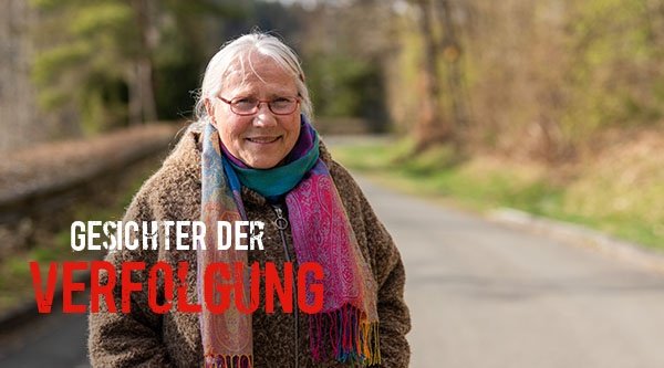 Eine Frau mit weißen Haaren steht im Wald und lächelt in die Kamera