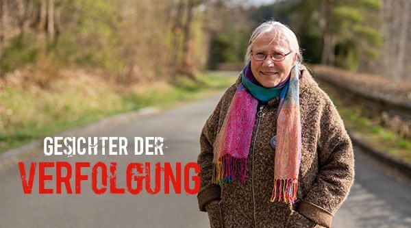 Eine Frau mit weißen Haaren steht im Wald und lächelt in die Kamera