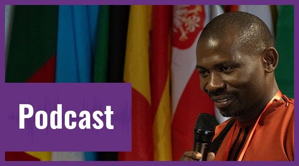 Ein Mann mit dunkler Hautfarbe im Close-up vor Flaggen fotografiert. Davor der Schriftzug "Podcast" auf lila Hintergrund.