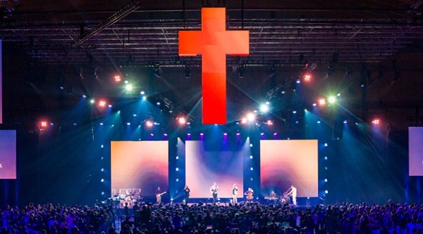 Ein riesiges beleuchtetes Kreuz schwebt über Tausenden Menschen in einer Messehalle