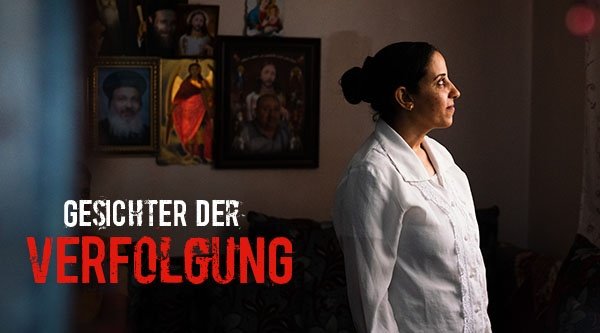 Gesichter der Verfolgung | Eine Frau steht in einer abgedunkelten Wohnung vor einem Fenster und wird vom Licht angestrahlt
