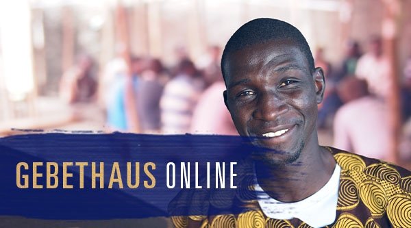 Ein afrikanischer Christ lächelt in Richtung des Betrachters. Im Hintergrund sitzen einige Menschen auf Bänken an Tischen