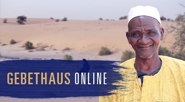 Ein Mann aus Mali lächelt vor einer kargen Landschaft in Richtung des Betrachters. Im Hintergrund reitet jemand auf einem Kamel.