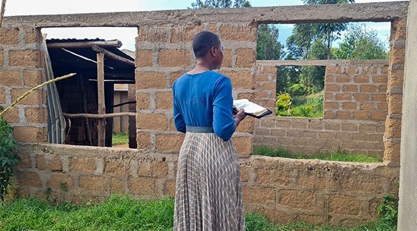 Eine afrikanische Frau mit einem blau-braunen Kleid steht vor einer einfachen Mauer aus Lehm und hält eine aufgeschlagene Bibel in der rechten Hand