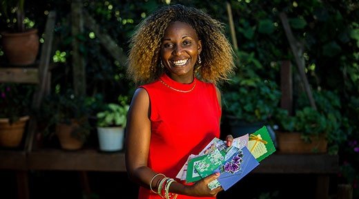 Eine afrikanische Frau in einem roten Kleid steht lächelnd vor grünen Büschen und Palmen und hält selbstgemachte Karten in den Händen