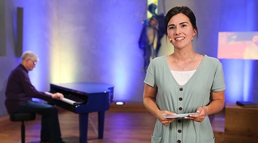 Eine junge Frau in einem hellgrünen Kleid spricht im Gebetshaus, während ein Mann im Hintergrund an einem Flügel sitzt und spielt