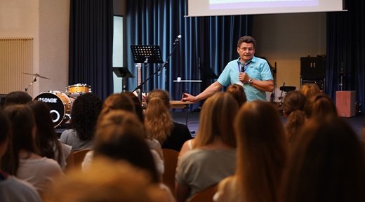 Ein Mann in blauem T-shirt steht vor der Bühne und spricht zu einer Gemeinde