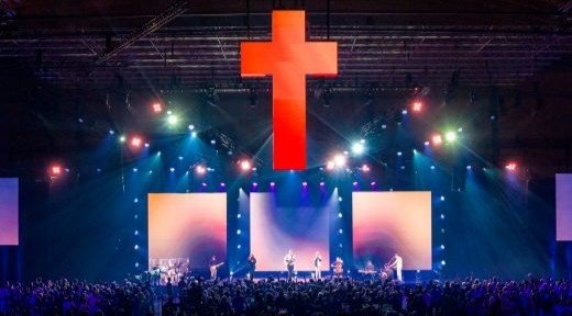 Tausende Menschen feiern Lobpreis unter einem riesigen beleuchteten Kreuz in einer Messehalle vor einer großen Bühne