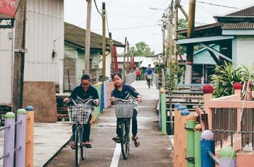 Zwei malaysische Mädchen fahren auf dem Fahrrad frontal auf die Kamera zu und hinter ihnen sieht man die von Häusern umgebene Gasse