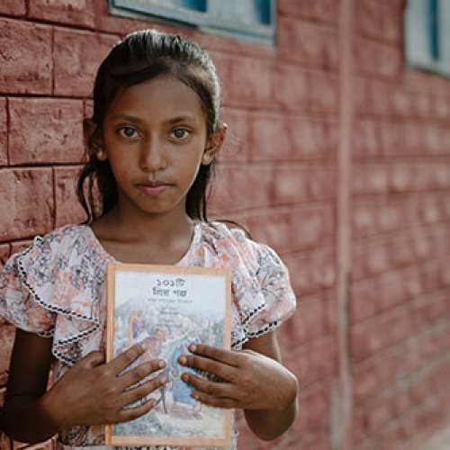 Ein Mädchen steht vor einem gemauerten Gebäude und hält ein Buch in der Hand.
