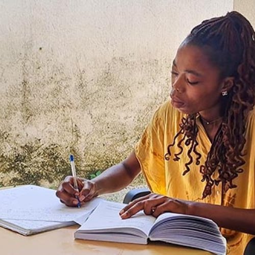 Eine afrikanische Frau macht sich Notizen zu einem aufgeschlagenem Buch. Sie trägt ein gelbes Oberteil.