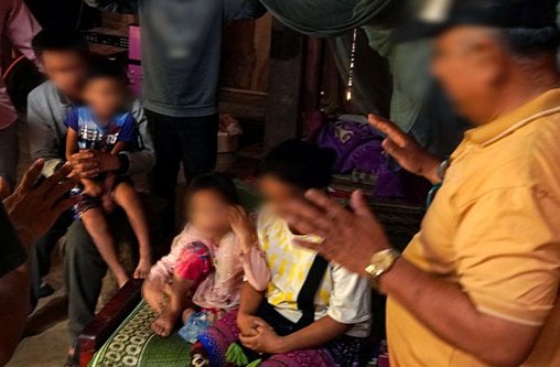 Kinder und Erwachsenen sitzen und ein Mann mit gelben Shirt hat seine Hände im Gebet ausgestreckt über die Kinder