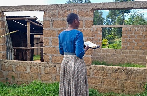 Eine afrikanische Frau mit einem blau-braunen Kleid steht vor einer einfachen Mauer aus Lehm und hält eine aufgeschlagene Bibel in der rechten Hand